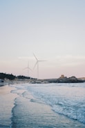 A serene coastal scene with gentle waves lapping at the shore. Two large wind turbines are prominently visible, standing against a soft sky. The horizon features a rugged coastline with scattered boulders, trees, and a few small structures. People are seen walking and enjoying the beach.