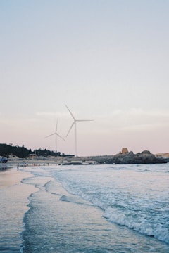 A serene coastal scene with gentle waves lapping at the shore. Two large wind turbines are prominently visible, standing against a soft sky. The horizon features a rugged coastline with scattered boulders, trees, and a few small structures. People are seen walking and enjoying the beach.