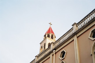 A church building's facade with a beige wall and small rectangular windows, featuring an ornate railing along the roofline. The steeple has a red roof topped with a cross, set against a clear blue sky.