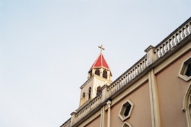 A church building's facade with a beige wall and small rectangular windows, featuring an ornate railing along the roofline. The steeple has a red roof topped with a cross, set against a clear blue sky.