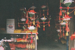 A shop interior filled with colorful, hanging decorations featuring floral designs and tassels. Below, various packaged goods and small golden figurines can be seen in a display case. The setting is dimly lit and presents a festive, vibrant atmosphere.