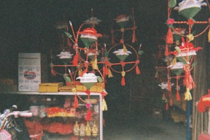 A shop interior filled with colorful, hanging decorations featuring floral designs and tassels. Below, various packaged goods and small golden figurines can be seen in a display case. The setting is dimly lit and presents a festive, vibrant atmosphere.