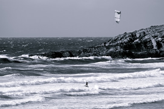 A windswept ocean scene with choppy waves crashing onto the shore. A lone kiteboarder is navigating the waves, using a large kite to harness the wind. A rocky cliff is visible in the background under a cloudy sky.