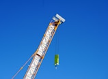 A crane arm extends diagonally across the image against a clear blue sky. The crane is yellow and white, with a hook and block attached by cables.