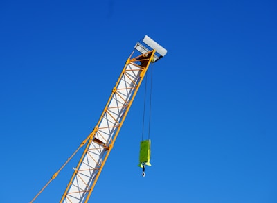 A crane arm extends diagonally across the image against a clear blue sky. The crane is yellow and white, with a hook and block attached by cables.