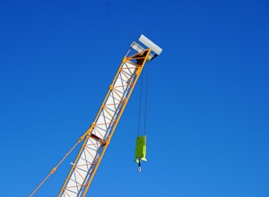 A crane arm extends diagonally across the image against a clear blue sky. The crane is yellow and white, with a hook and block attached by cables.