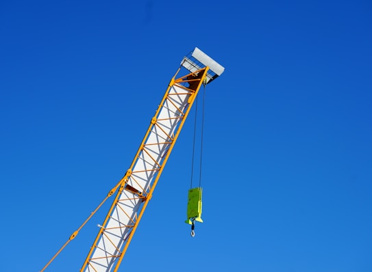 A crane arm extends diagonally across the image against a clear blue sky. The crane is yellow and white, with a hook and block attached by cables.