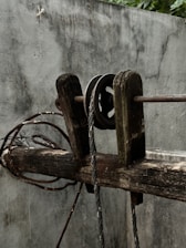 A weathered wooden pulley system is set against a rough grey wall. The pulley is attached to a thick, frayed rope and appears worn with age. Coiled wires are visible beside the wooden beam, adding to the rustic and aged appearance of the scene.