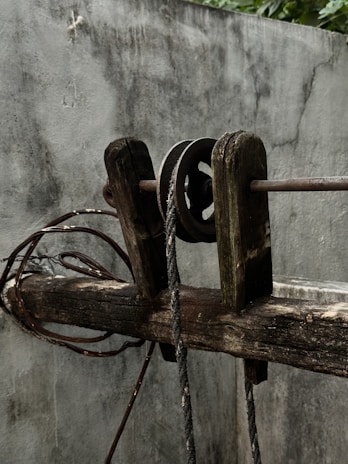 A weathered wooden pulley system is set against a rough grey wall. The pulley is attached to a thick, frayed rope and appears worn with age. Coiled wires are visible beside the wooden beam, adding to the rustic and aged appearance of the scene.