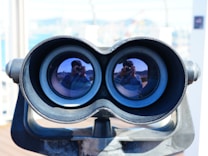 A pair of large binoculars with reflections of a person taking a photo, visible in both lenses. The binoculars are mounted on a stand and have a weathered appearance.
