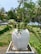 A large white ceramic pot is placed on a stone pedestal surrounded by lush greenery. The pot contains a plant with broad, slightly curved green leaves. In the background, there are various types of trees, including palm trees, suggesting a tropical environment.