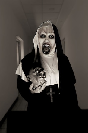 A person dressed in a nun's habit with eerie makeup, showing dark circles around the eyes, is standing in a dimly lit hallway. The person is holding a severed mannequin head with grotesque features and has a large cross hanging around their neck.