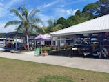 A marina or watersports facility with a variety of boats and equipment organized in racks. Several canoes and kayaks are visible. Palm trees and lush greenery surround the open-air structure, providing a tropical setting. A purple tent canopy and a sign reading 'Open to Public' are prominently displayed.