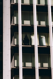 A portion of a modern glass building facade featuring large windows framed by vertical and horizontal metal beams. Sunlight casts shadows on the reflective surfaces, with one window displaying partially opened curtains.