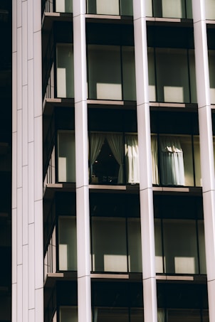 A portion of a modern glass building facade featuring large windows framed by vertical and horizontal metal beams. Sunlight casts shadows on the reflective surfaces, with one window displaying partially opened curtains.