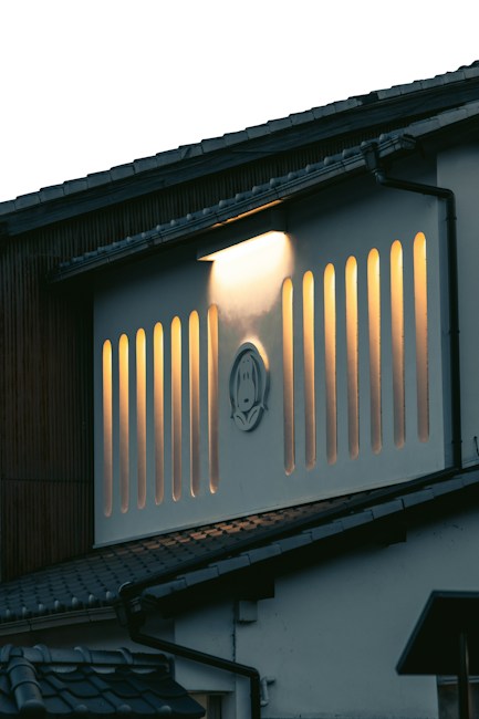 A building features vertical slits backlit with a warm glow, creating an artistic pattern on the fa&ccedil;ade. A unique emblem is positioned at the center of these slits. The structure is topped with a tiled roof, and the lighting conveys a sense of calm during twilight.
