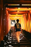 A person wearing a Neymar Jr. football jersey walks up a stairway lined with vibrant orange torii gates, creating a tunnel-like effect with sunlight filtering through the structure. Shadows and light interplay, creating a sense of depth and perspective.