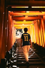 A person wearing a Neymar Jr. football jersey walks up a stairway lined with vibrant orange torii gates, creating a tunnel-like effect with sunlight filtering through the structure. Shadows and light interplay, creating a sense of depth and perspective.