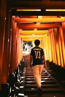 A person wearing a Neymar Jr. football jersey walks up a stairway lined with vibrant orange torii gates, creating a tunnel-like effect with sunlight filtering through the structure. Shadows and light interplay, creating a sense of depth and perspective.