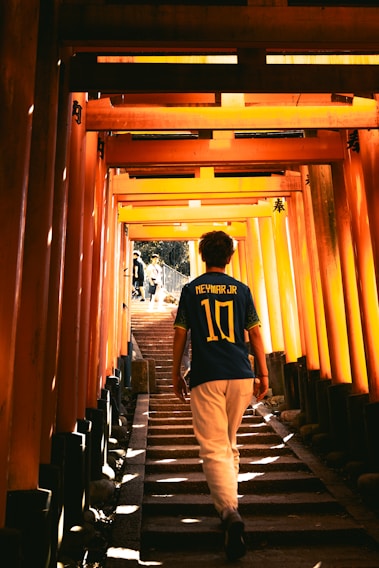 A person wearing a Neymar Jr. football jersey walks up a stairway lined with vibrant orange torii gates, creating a tunnel-like effect with sunlight filtering through the structure. Shadows and light interplay, creating a sense of depth and perspective.