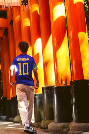 A person wearing a sports jersey with the name and number 'Neymar Jr 10' walks through a passageway lined with tall, vibrant orange pillars. The pillars are partially shaded and have some text written on them, likely in Japanese characters. The ground is paved, and the scene is illuminated by warm sunlight.