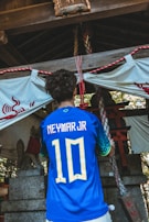 A person is wearing a blue soccer jersey with 'Neymar Jr' and the number '10' on the back, standing in front of a traditional shrine. The shrine features stone statues and rope decorations, with a white cloth adorned with red symbols.