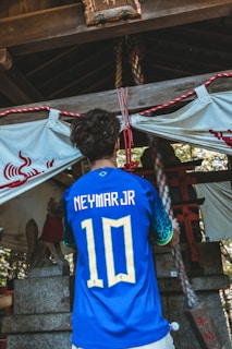 A person is wearing a blue soccer jersey with 'Neymar Jr' and the number '10' on the back, standing in front of a traditional shrine. The shrine features stone statues and rope decorations, with a white cloth adorned with red symbols.