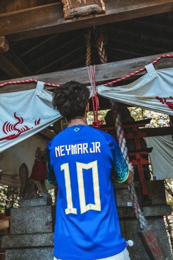 A person is wearing a blue soccer jersey with 'Neymar Jr' and the number '10' on the back, standing in front of a traditional shrine. The shrine features stone statues and rope decorations, with a white cloth adorned with red symbols.