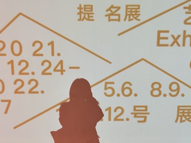 Silhouette of a person standing in front of a light background displaying text in multiple languages, including dates and terms related to exhibitions. The text is written in bold, prominent letters in orange color.