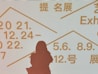 Silhouette of a person standing in front of a light background displaying text in multiple languages, including dates and terms related to exhibitions. The text is written in bold, prominent letters in orange color.