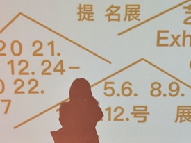 Silhouette of a person standing in front of a light background displaying text in multiple languages, including dates and terms related to exhibitions. The text is written in bold, prominent letters in orange color.