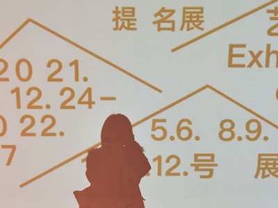 Silhouette of a person standing in front of a light background displaying text in multiple languages, including dates and terms related to exhibitions. The text is written in bold, prominent letters in orange color.