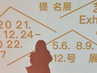 Silhouette of a person standing in front of a light background displaying text in multiple languages, including dates and terms related to exhibitions. The text is written in bold, prominent letters in orange color.