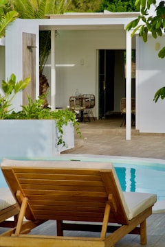 A serene outdoor patio featuring a wooden lounge chair by a clear blue pool. The area is surrounded by lush greenery and partially enclosed by a modern white building with a pergola. Inside, there are wicker chairs and a table visible through the open entrance.