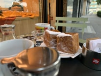 A close-up view of a table set outside a cafe, featuring a plate with two pieces of pastry dusted with powdered sugar. Behind them are several cups, including a white mug and a small glass with water. To the left, there is a bottle with a metal cap and a cream dispenser. The background shows a window display with a fabric draped over a counter, and part of the street can be seen on the right.