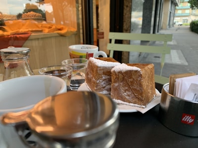 A close-up view of a table set outside a cafe, featuring a plate with two pieces of pastry dusted with powdered sugar. Behind them are several cups, including a white mug and a small glass with water. To the left, there is a bottle with a metal cap and a cream dispenser. The background shows a window display with a fabric draped over a counter, and part of the street can be seen on the right.