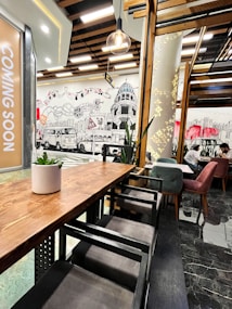 A modern cafe interior with wooden furniture and artistic wall murals depicting cityscape elements. The space features a wooden ceiling with hanging light fixtures. Comfortable seating with velvet chairs in shades of green and red can be seen, and a plant decorates a wooden table. A 'Coming Soon' sign is visible on the left.
