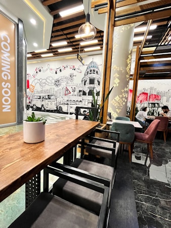 A modern cafe interior with wooden furniture and artistic wall murals depicting cityscape elements. The space features a wooden ceiling with hanging light fixtures. Comfortable seating with velvet chairs in shades of green and red can be seen, and a plant decorates a wooden table. A 'Coming Soon' sign is visible on the left.