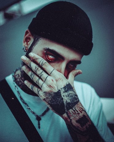 A person wearing a dark beanie covers their mouth with a tattooed hand. The hand features intricate designs, including a large butterfly tattoo. The person has red eyes and a piercing in the eyebrow. The atmosphere is dimly lit, adding a mysterious and intense vibe.