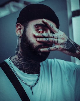 A person with extensive tattoos on the neck, hand, and face is partially covering their face with their hand, revealing a piercing and an exposed eye. The person is wearing a black beanie, earrings, and a chain necklace. They have a serious expression and the overall tone is dark and moody.