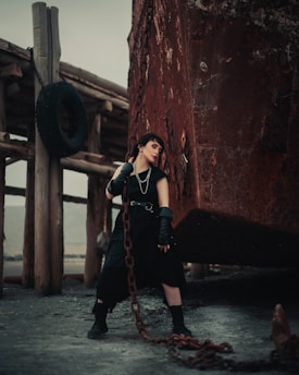 A person dressed in dark, edgy clothing leans against a rusty, corroded metal structure with a thick chain. The background consists of wooden supports and an old tire hanging, creating a rugged, industrial setting.