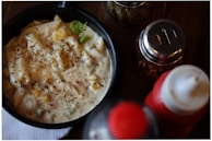 A black bowl filled with creamy pasta sauce, topped with herbs, corn, and bits of seasoning, accompanied by a green garnish leaf on wood table. Adjacent to the bowl are condiment containers, including a red and white bottle and spice jars.
