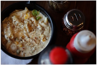 A black bowl filled with creamy pasta sauce, topped with herbs, corn, and bits of seasoning, accompanied by a green garnish leaf on wood table. Adjacent to the bowl are condiment containers, including a red and white bottle and spice jars.