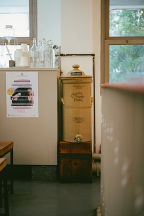 A vintage-style water filter with a classic design stands next to a counter. The counter is topped with glass bottles and stacked paper cups. A colorful poster is visible on the side of the counter. The room features large windows with a view of greenery outside.