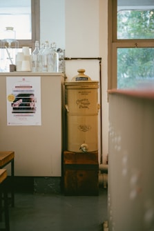 A vintage-style water filter with a classic design stands next to a counter. The counter is topped with glass bottles and stacked paper cups. A colorful poster is visible on the side of the counter. The room features large windows with a view of greenery outside.