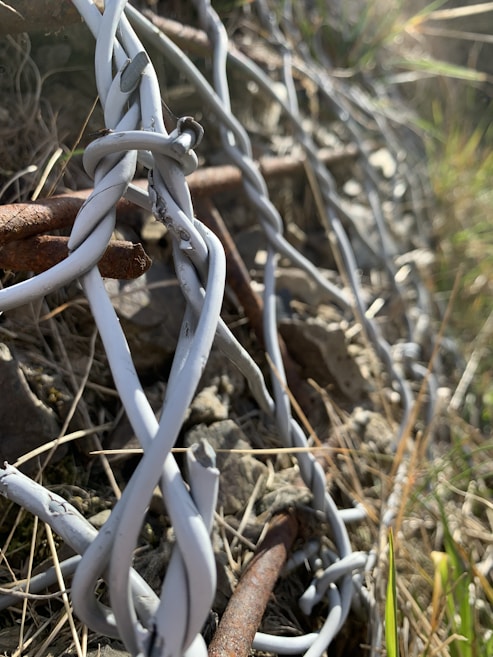 Close-up of intertwined metal wires, primarily grey, coiled around a rusty rod. The background is a textured mixture of rock and dry grass, with sunlight casting subtle shadows on the wires.