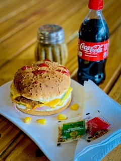 A delicious burger with a sesame seed bun, layered with cheese, lettuce, and a patty is placed on a white plate. Next to the plate is a bottle of Coca-Cola and a small shaker, possibly containing spices. Ketchup is drizzled on top of the bun, and a few corn kernels are scattered around. Two packets, one labeled 'Oregano Spice Sprinkler' and another 'Hot Chili Flakes', rest alongside a folded napkin.