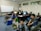 A group of six people are gathered in a computer room, some sitting on the floor and others on chairs. They are casually dressed and appear to be engaged in a relaxed activity, with one person flashing a peace sign and another smiling brightly. The room contains several computers on desks and a large whiteboard in the background with some writing on it.