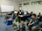 A group of six people are gathered in a computer room, some sitting on the floor and others on chairs. They are casually dressed and appear to be engaged in a relaxed activity, with one person flashing a peace sign and another smiling brightly. The room contains several computers on desks and a large whiteboard in the background with some writing on it.