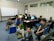 A group of six people are gathered in a computer room, some sitting on the floor and others on chairs. They are casually dressed and appear to be engaged in a relaxed activity, with one person flashing a peace sign and another smiling brightly. The room contains several computers on desks and a large whiteboard in the background with some writing on it.